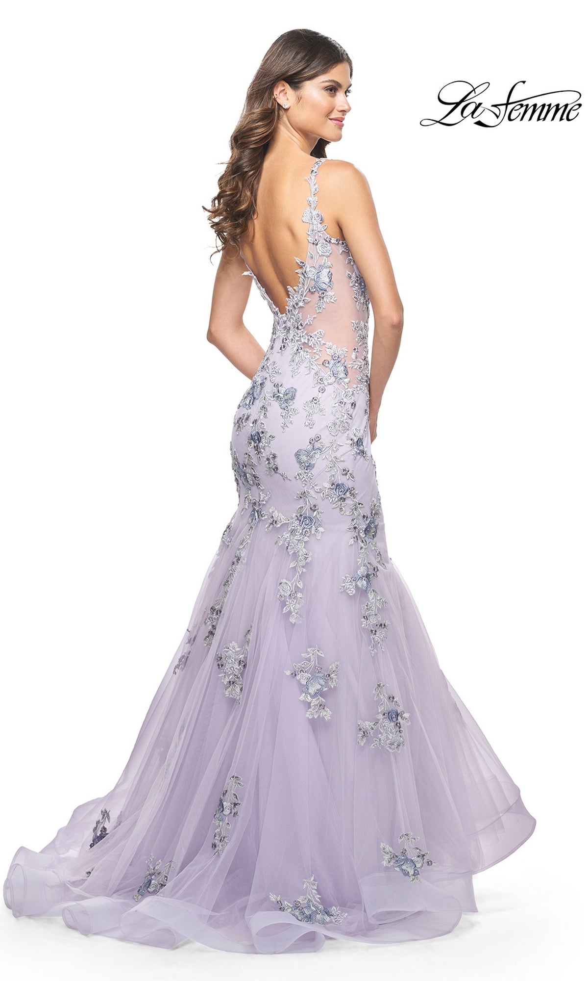 A woman wearing the La Femme Long Lavender Mermaid Prom Dress 32091, featuring floral appliqué details and an open back, poses with her back slightly turned.